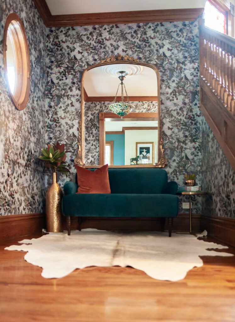 Eclectic entryway with dramatic wallpaper, teal velvet loveseat, cowhide rug, and vintage mirror.