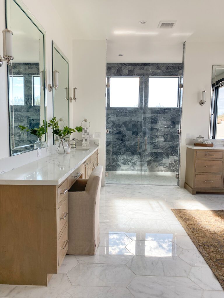 Bright luxury bathroom with warm wood vanity, marble flooring, expansive mirror, and natural light.