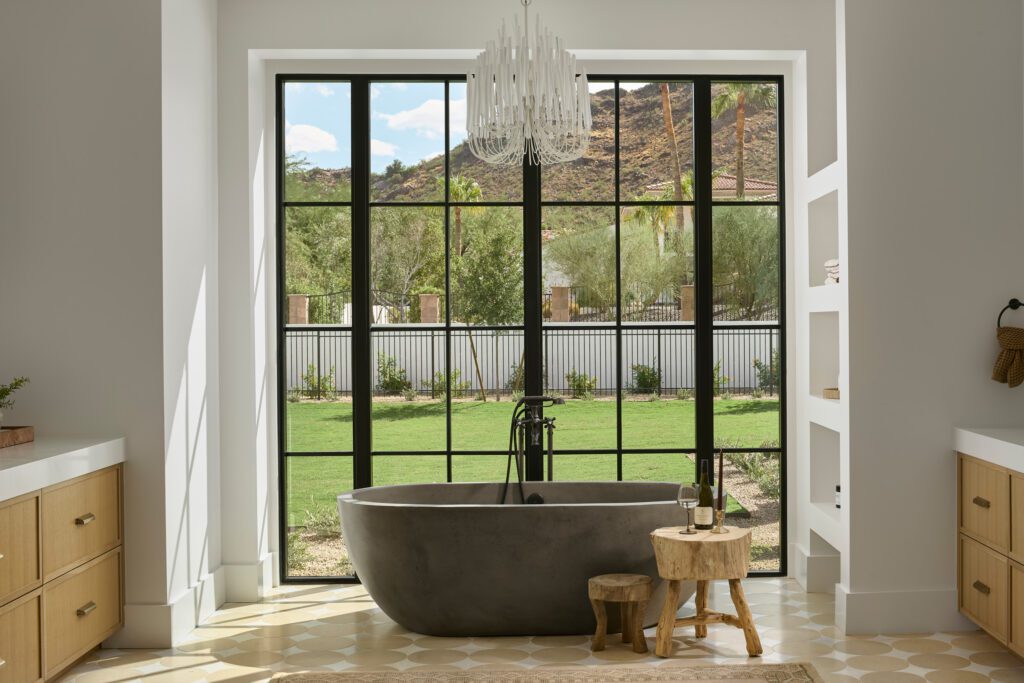 Modern freestanding bathtub beneath a sculptural chandelier with natural light pouring through black steel frame windows in a luxury Paradise Valley home.