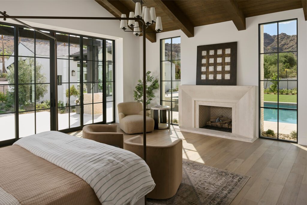 Luxury bedroom with exposed wood beams, modern fireplace, neutral furnishings, and warm layered textures.