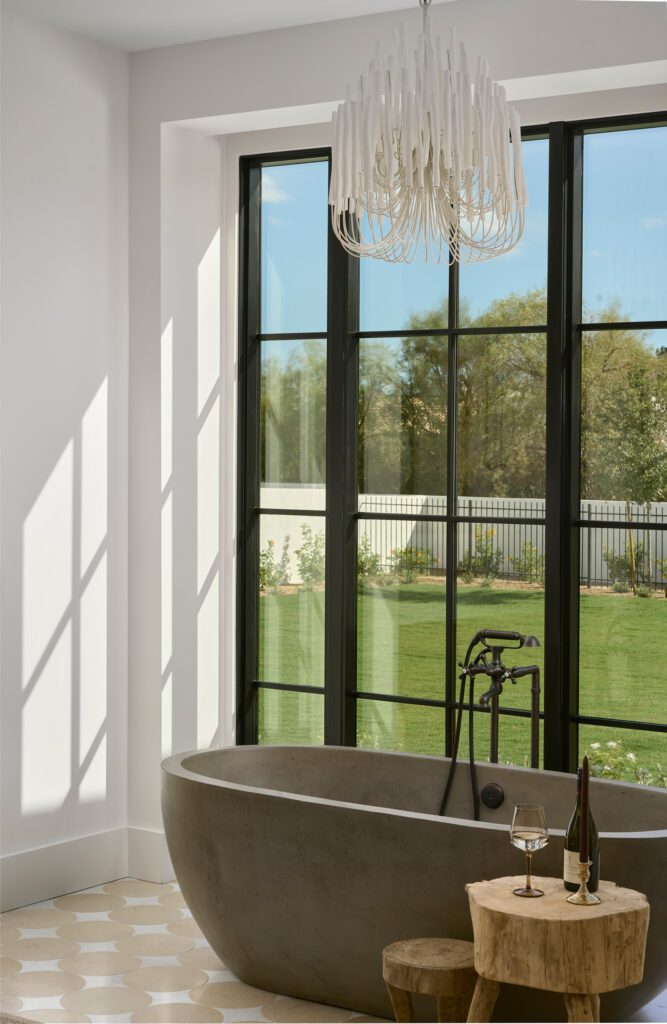 A luxurious bathroom with a freestanding bathtub positioned in front of a massive black-framed window overlooking a green outdoor space, offering a natural spa experience.