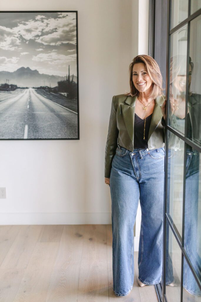 Meredith, the luxury interior designer for The Smyth House, stands confidently in a stylish olive jacket and jeans, beside a large framed desert landscape print and a black-framed glass door.