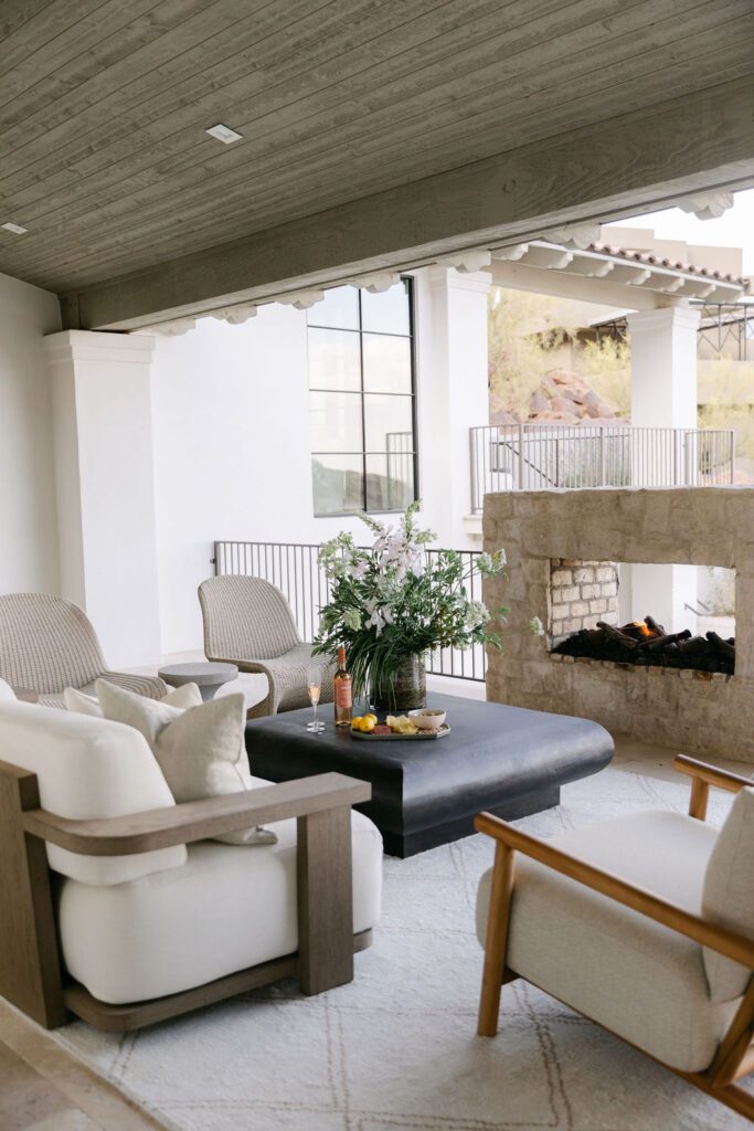 A luxurious outdoor living room featuring a grand stone fireplace, elegant seating, and a coffee table, integrated with a modern home's interior via large openings.