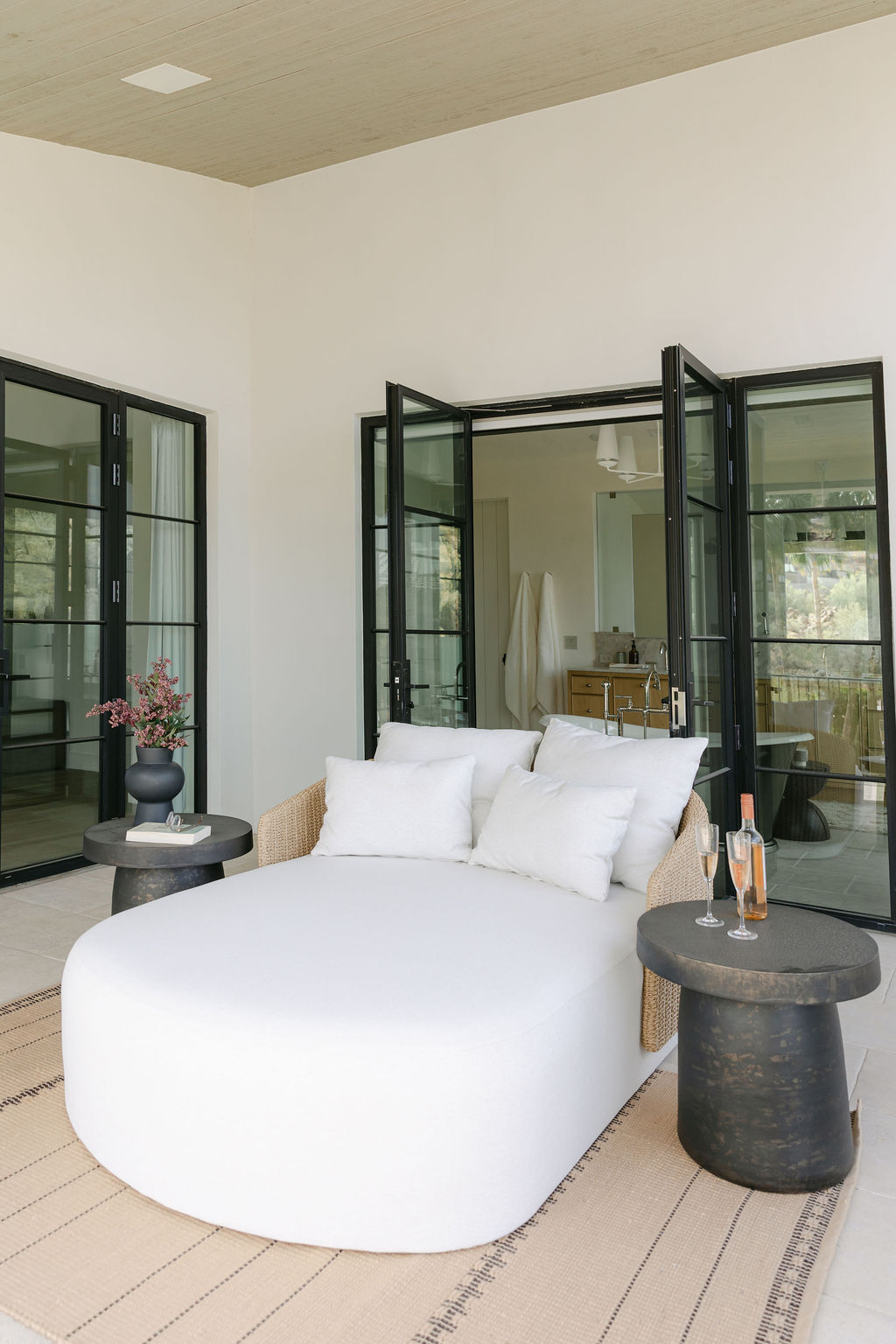 A luxurious outdoor daybed on a light-colored rug, positioned on a patio leading to a bright interior through black-framed bi-fold glass doors, showcasing seamless indoor-outdoor living.