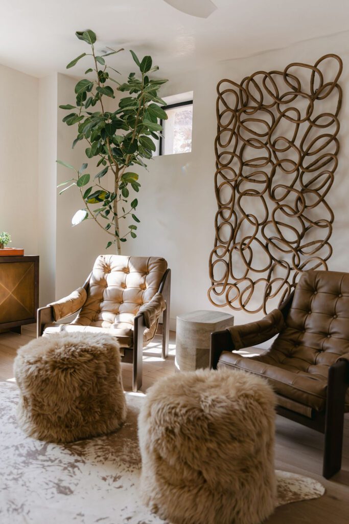 Earth-toned lounge with tufted leather chairs, wooly stools, and abstract wood wall sculpture.