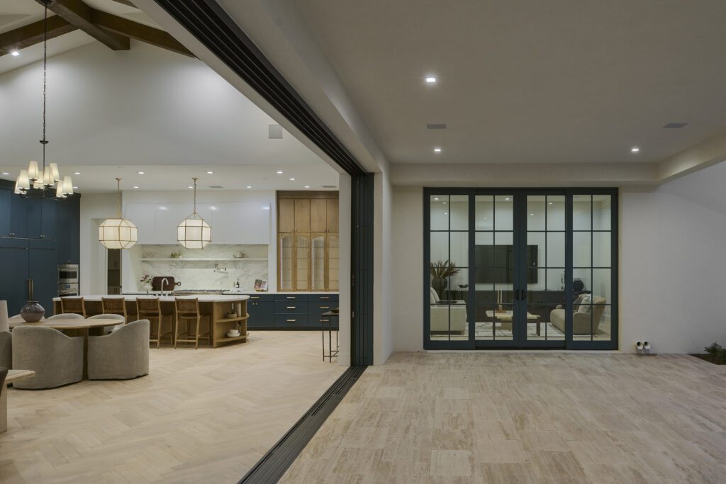 A wide-angle view of a modern open-concept luxury home in Phoenix, featuring a teal and white kitchen and an expansive opening to a large, versatile living space with light tile flooring.