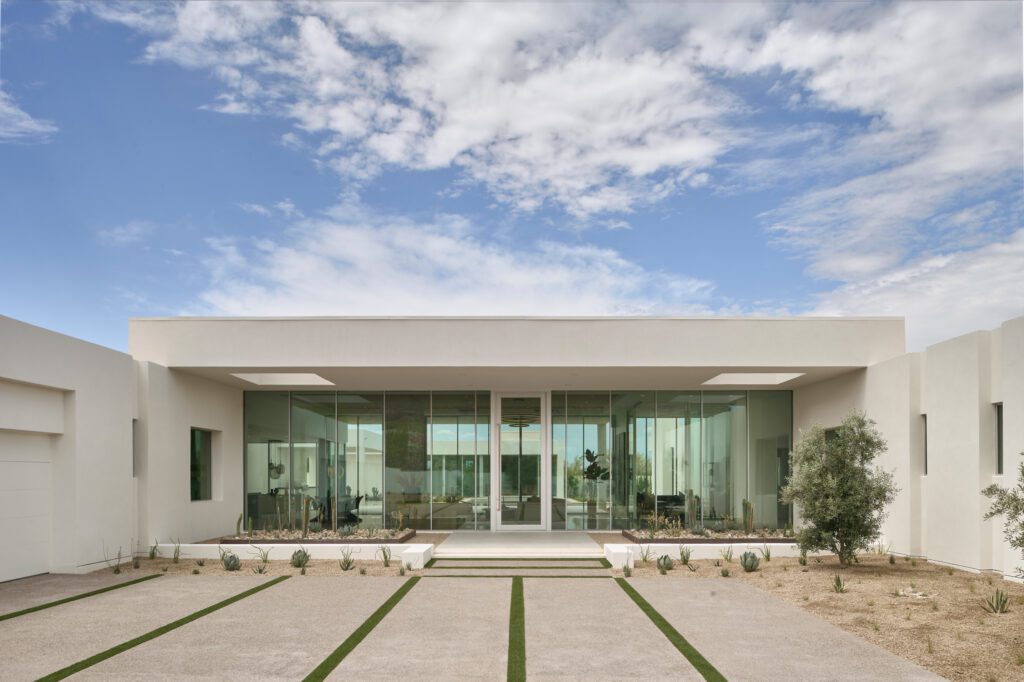 The striking front elevation of a modern luxury home in Phoenix, featuring a prominent glass facade that offers transparency into the interior, surrounded by minimalist desert landscaping and a concrete paver pathway.