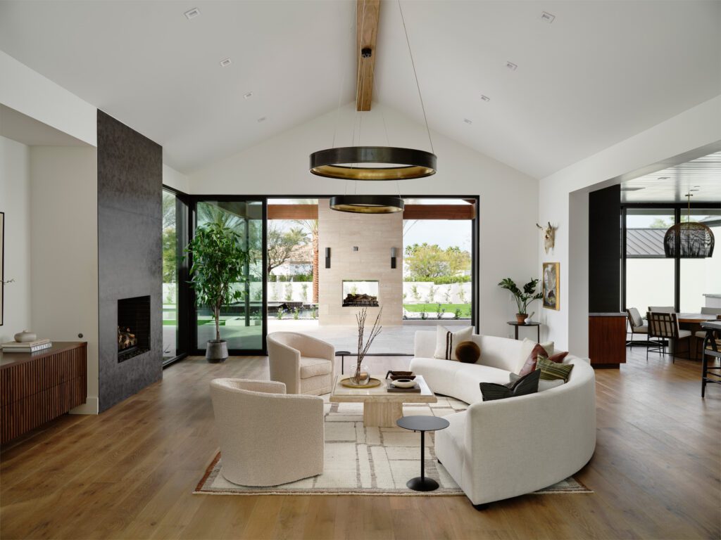 A luxurious open-concept living room with a modern fireplace and curved sofa, featuring expansive sliding glass doors that fully open to an outdoor patio with a pool and another fireplace, embodying seamless indoor-outdoor design in Phoenix.