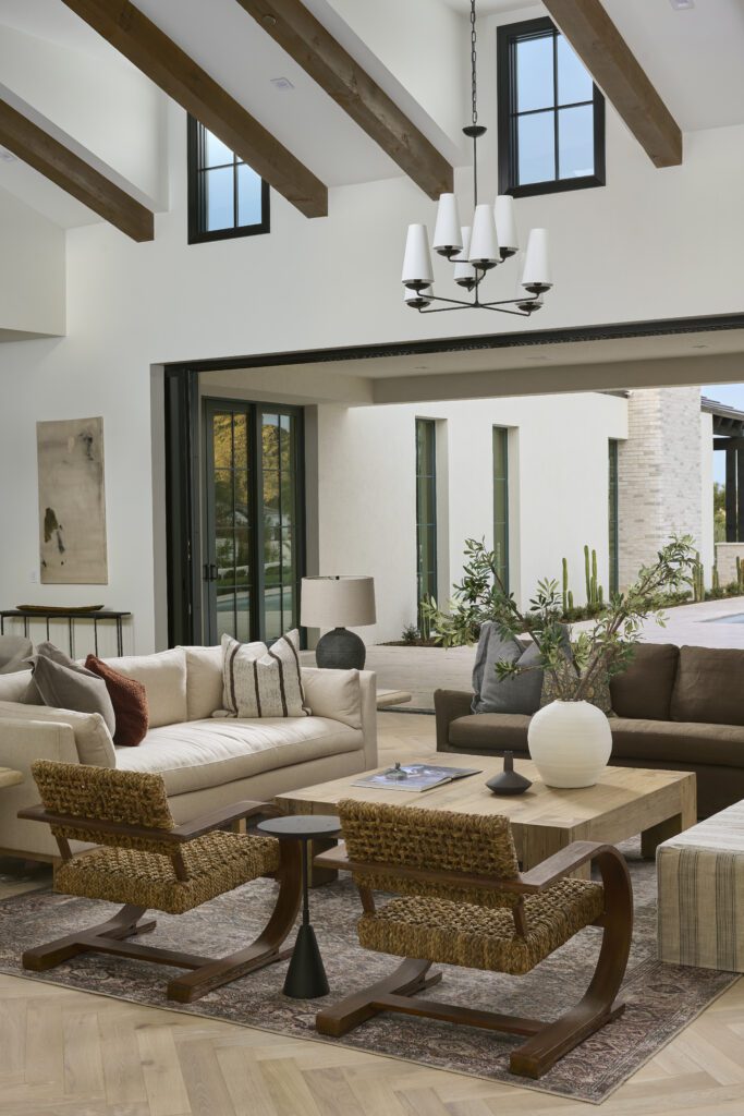 A luxurious living room with vaulted wooden beam ceilings and a chandelier, opening widely to an outdoor patio with desert landscaping, showcasing seamless indoor-outdoor design.