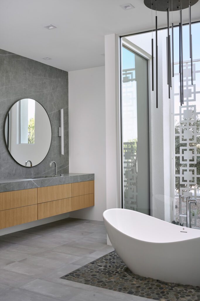 Modern luxury bathroom with floating wood vanity, round mirror, concrete walls, and freestanding tub.