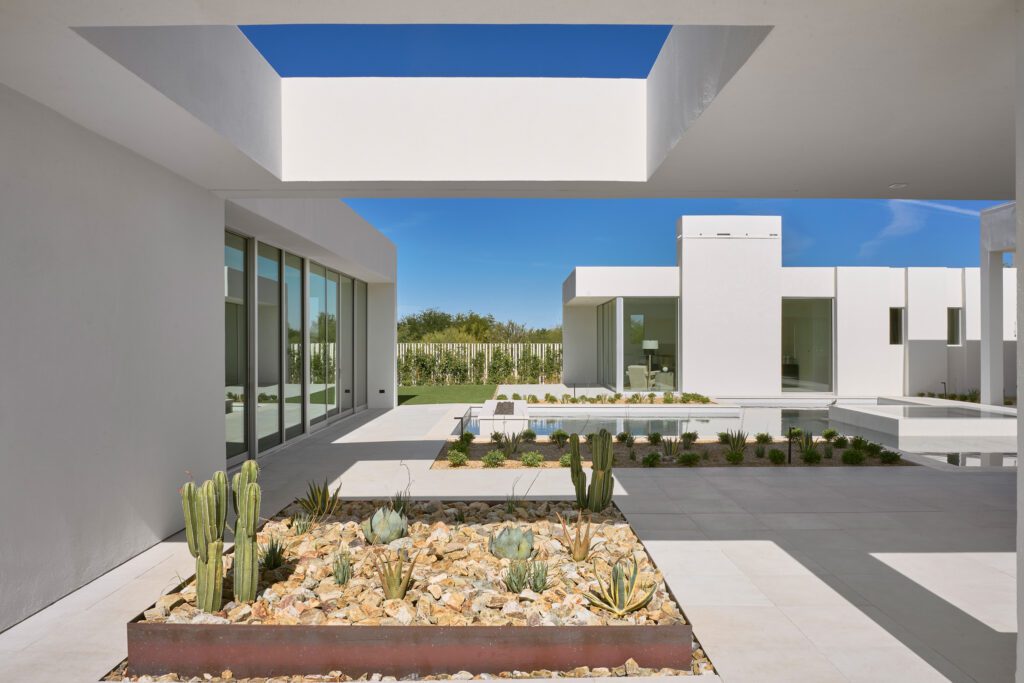 A modern white home exterior with expansive glass doors opening to a minimalist desert courtyard featuring gravel, cacti, and a unique overhead architectural opening, signifying seamless entry.