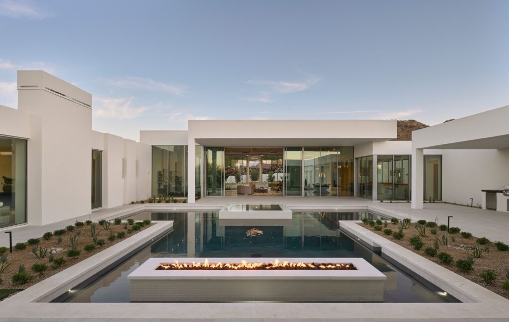 A wide exterior view of a modern luxury home in Phoenix with an expansive pool, an integrated linear fire pit, and large glass openings connecting interior and exterior spaces.