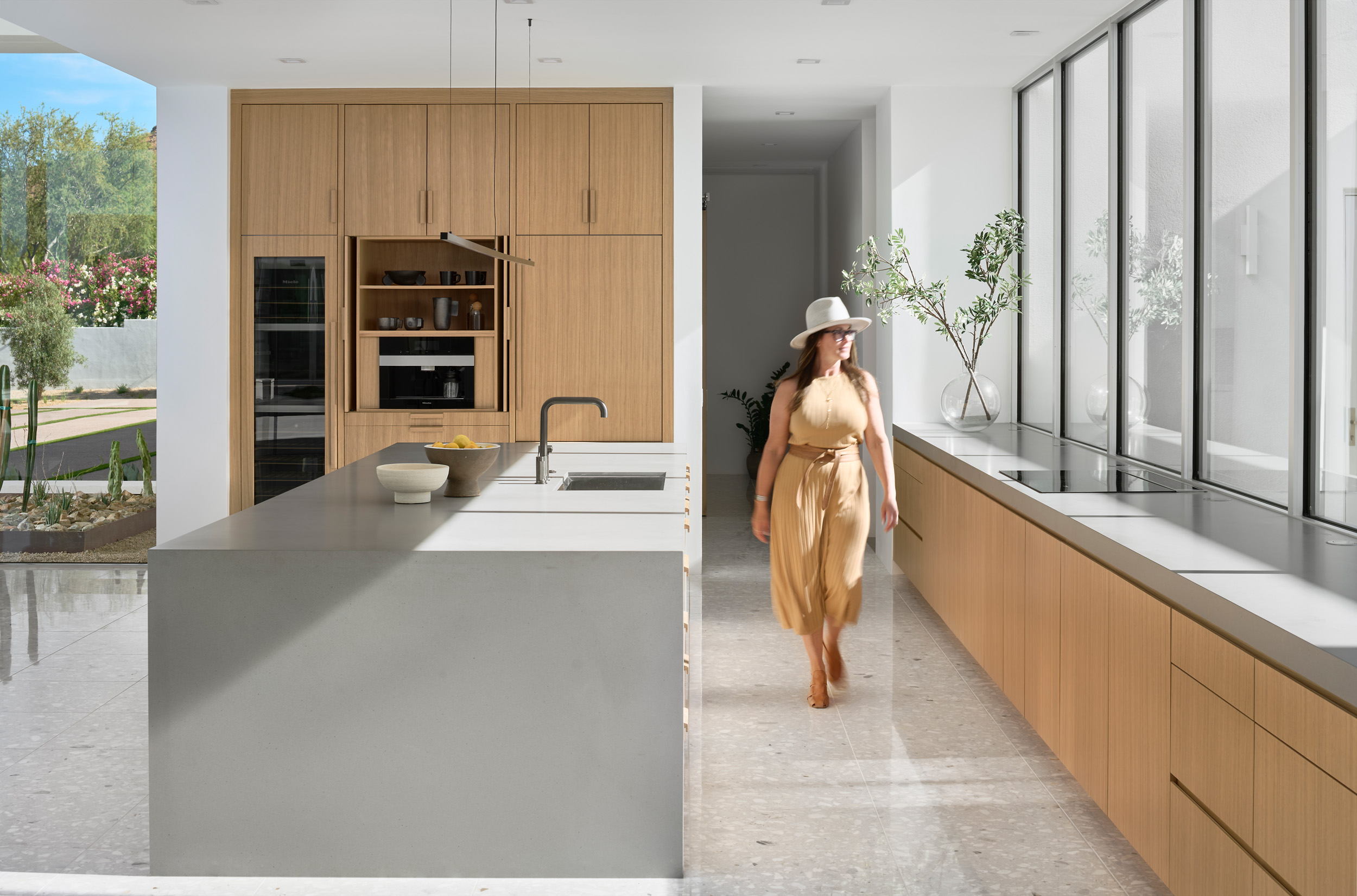 Luxury desert kitchen with minimalist design, light oak cabinetry, concrete island, and designer walking through a sunlit space in an Arizona home.