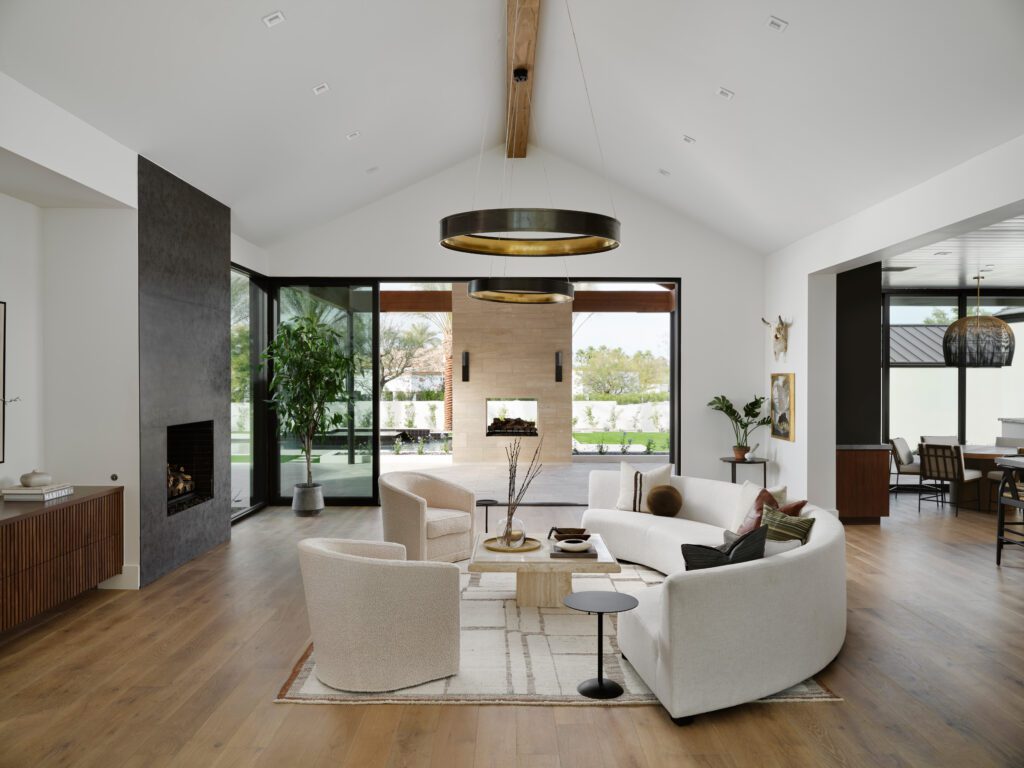 Modern desert living room with curved white sofa, boucle chairs, and natural light opening to a fireplace and outdoor space.
