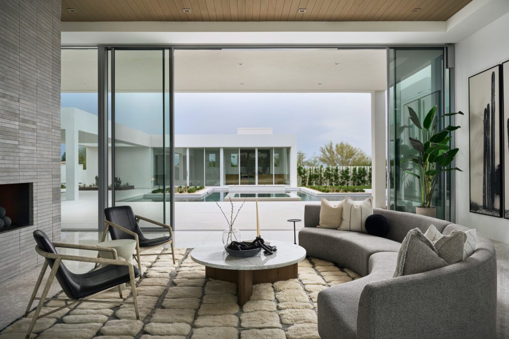 Modern luxury living room with curved gray sofa, stone fireplace, and sliding glass doors opening to a pool courtyard in a desert home designed for entertaining.
