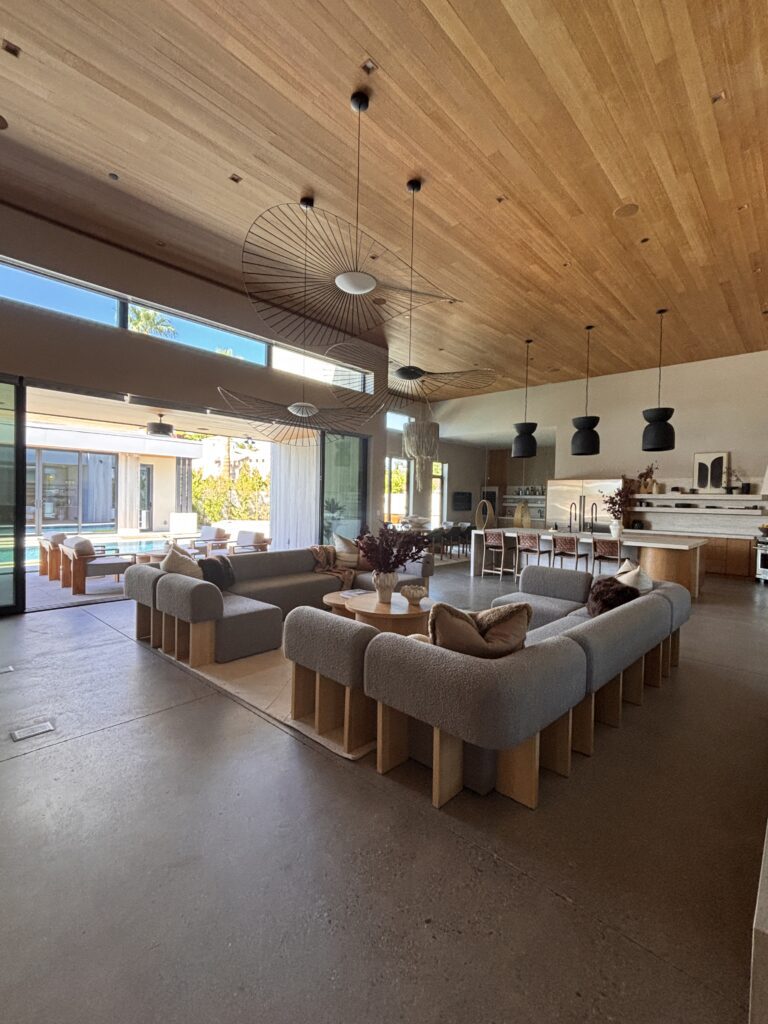 A spacious open-concept living and kitchen area with warm wood ceilings, neutral furnishings, sculptural lighting, and large sliding doors opening to the outdoor space.