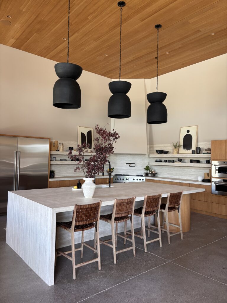 A modern kitchen with a long stone island, warm wood cabinetry, sculptural black pendant lights, open shelving, and neutral desert-toned styling.