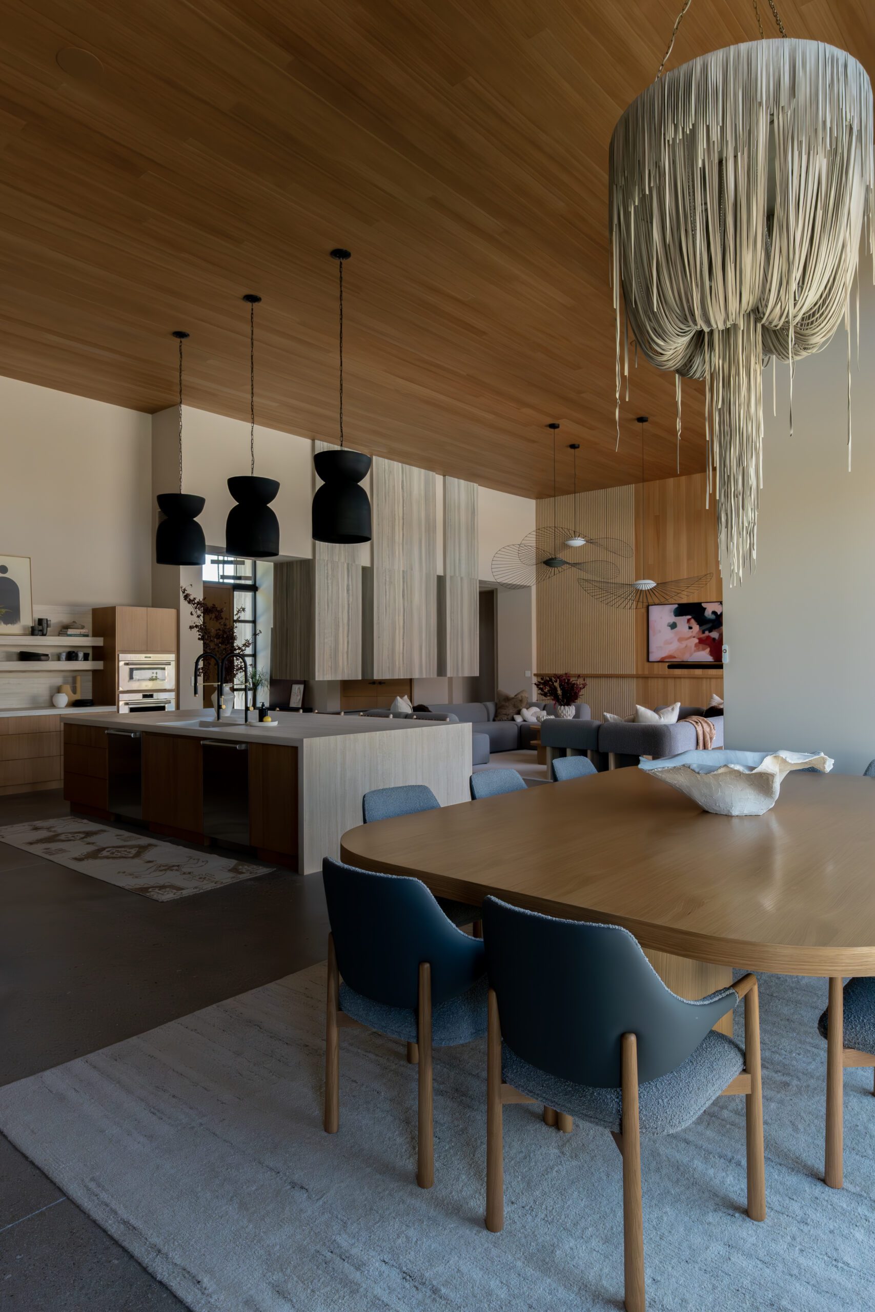 Open-concept living and dining space featuring dramatic sculptural lighting, including oversized black pendants and a cascading fringe chandelier, paired with warm wood ceilings and modern furnishings.