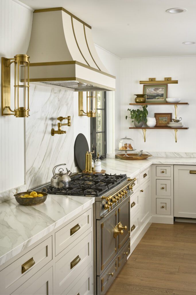 Custom cream and brass range hood in a luxury kitchen with marble backsplash, brass sconces, and a high-end French range shapes what 2026 interior design trends are all about.