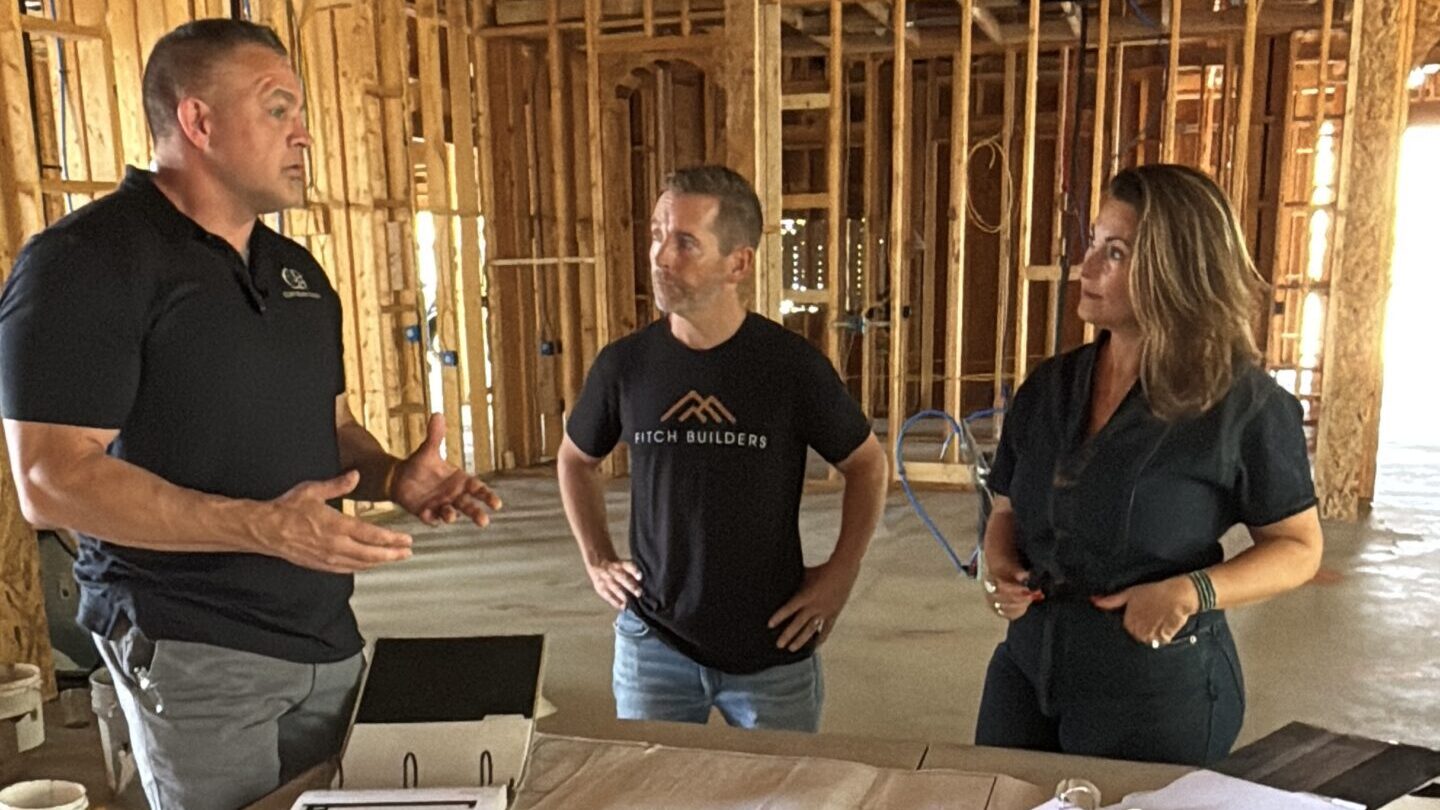 Three experts, the designer, builder, and architect, review plans on the framed construction site.