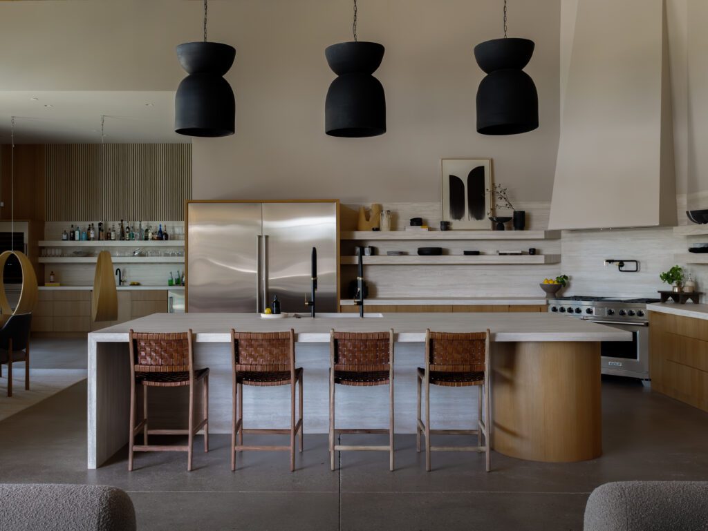 Modern kitchen interior featuring sculptural black clay pendant lights over a stone island, layered wood cabinetry, open shelving, and woven bar stools, styled in a warm, minimalist luxury aesthetic.
