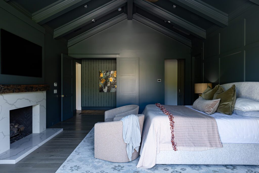 Moody luxury bedroom with dark blue walls, layered neutral bedding, modern lighting, and soft textural accents.