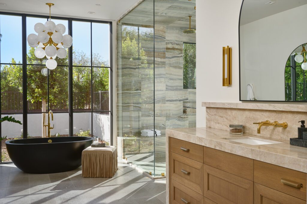 Bright modern bathroom with warm wood vanity, natural stone finishes, floor-to-ceiling glass, and indoor-outdoor views.