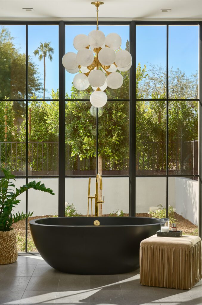 Luxury primary bathroom designed by Smyth House featuring a sculptural black soaking tub, dramatic floor-to-ceiling steel windows, brass plumbing fixtures, and statement chandelier in a modern Arizona home.
